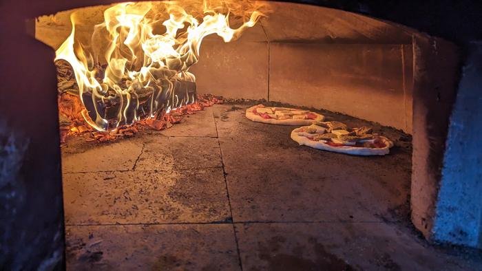 Two pizzas cooking on the oven floor beside the fire