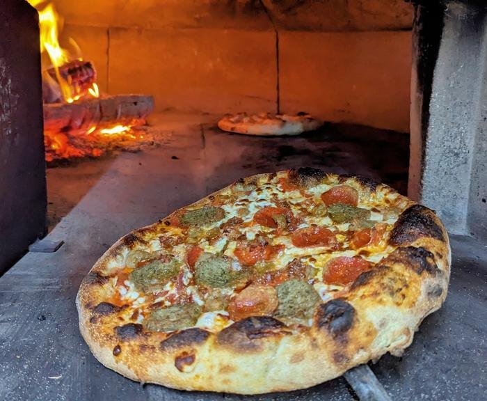 A pizza baking in the wood-fired oven with the flame behind it